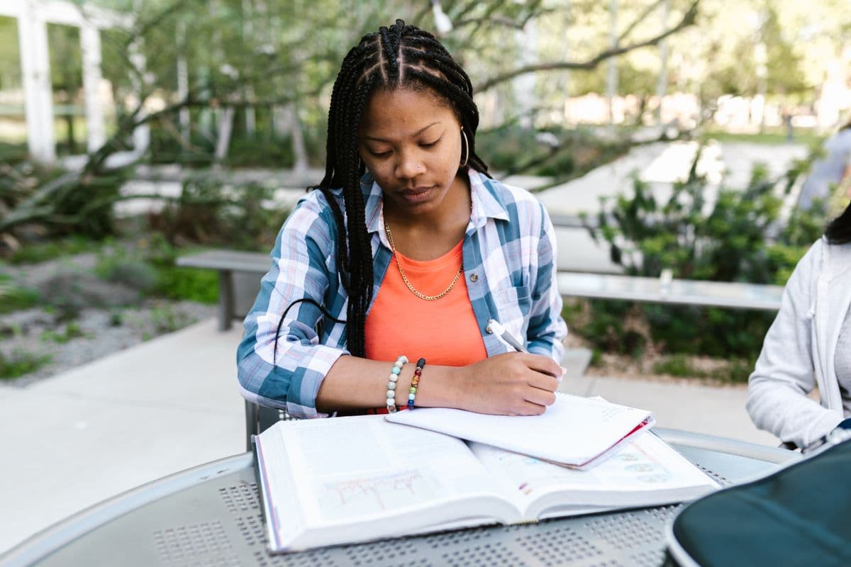 A student writing notes at a campus table beside an open textbook
